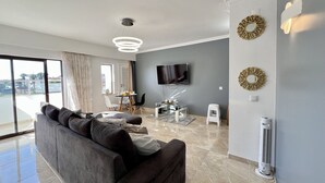 Una sala moderna con un sofá gris, un televisor de pantalla plana, espejos decorativos en la pared y un comedor con mesa y sillas.