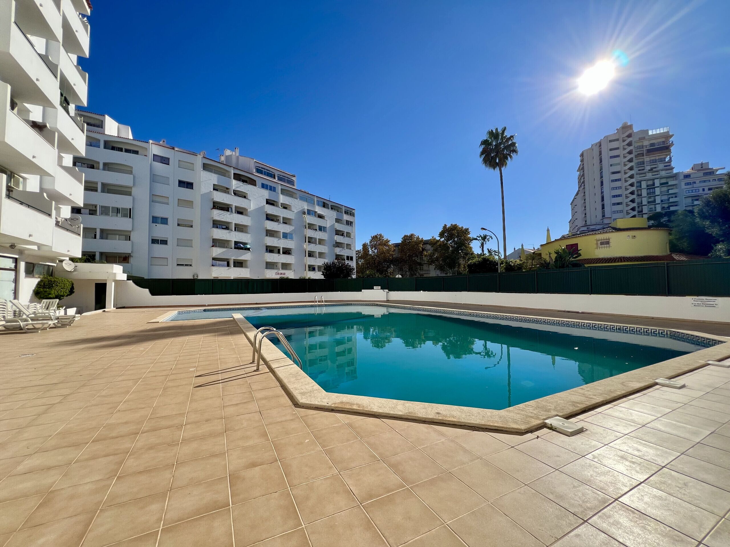 Une piscine au sol carrelé, un palmier et un immeuble à plusieurs étages en arrière-plan.