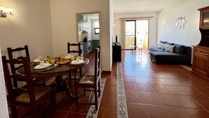 Zona pranzo con tavolo rotondo, sedie e prodotti per la colazione. Sullo sfondo, una cucina con pensili e lavello. In primo piano, si vede una zona living con divano e televisore.