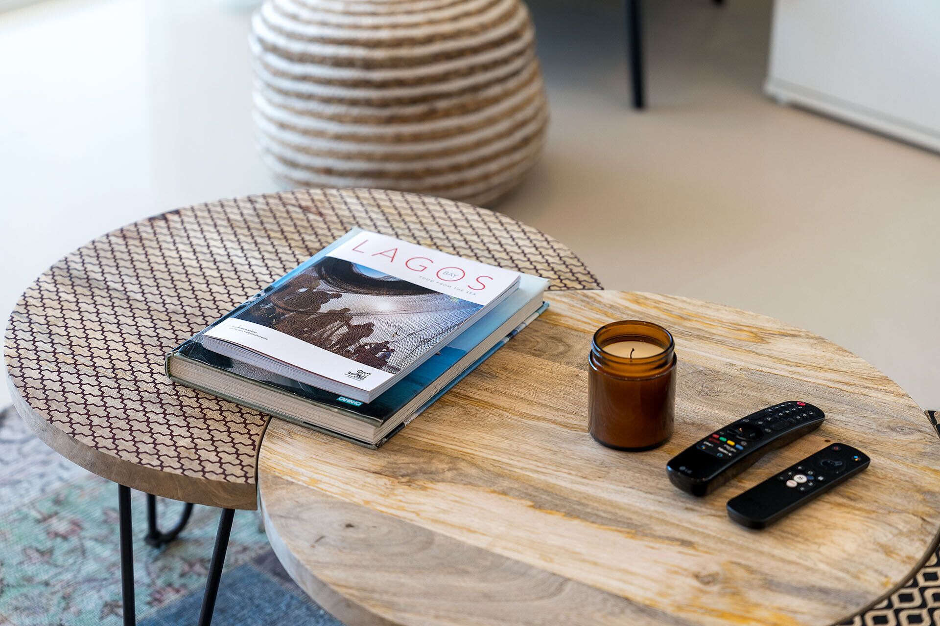 A round wooden table with a book titled 'Lagos', two remote controls, and a candle.