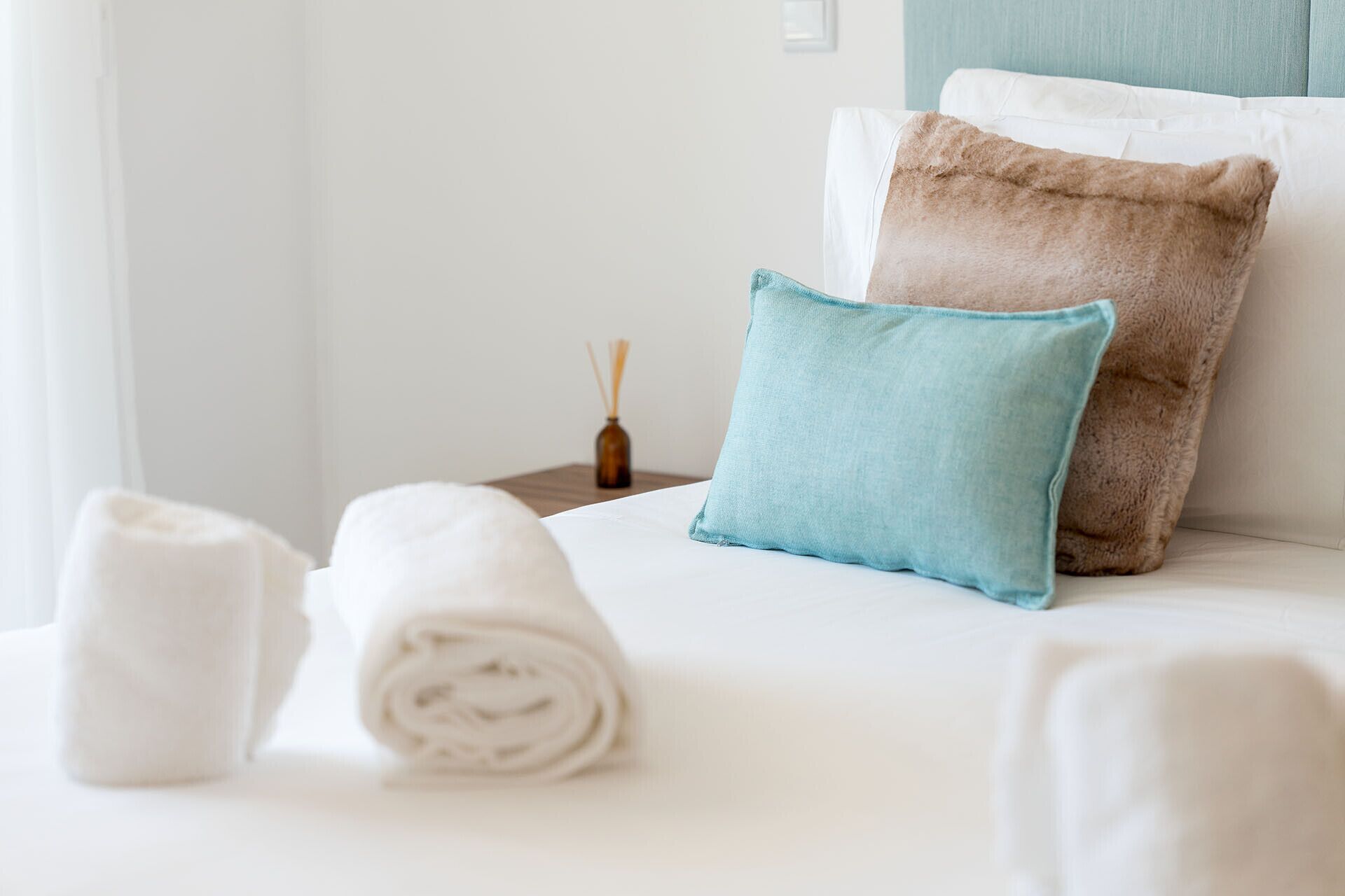 A neatly made bed with two rolled towels and two pillows, one light blue and one brown, in a hotel room with a simple, clean design.