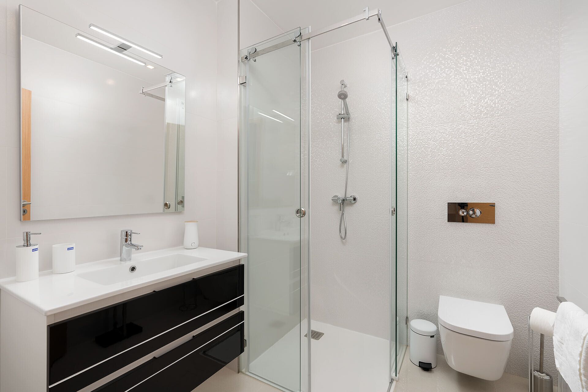 Un baño moderno con cabina de ducha de vidrio, lavamanos blanco sobre mueble negro y un inodoro.