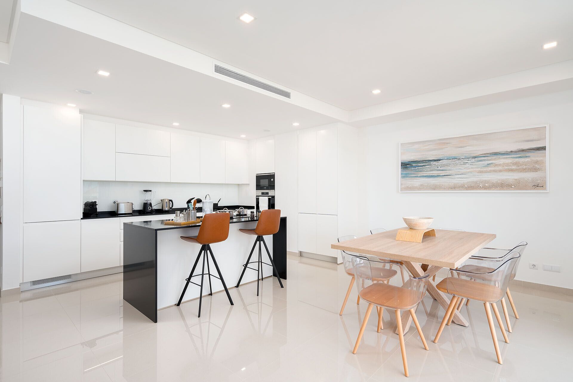 Une cuisine moderne avec des meubles blancs, un îlot central doté de tabourets et un coin repas avec une table et des chaises en bois.