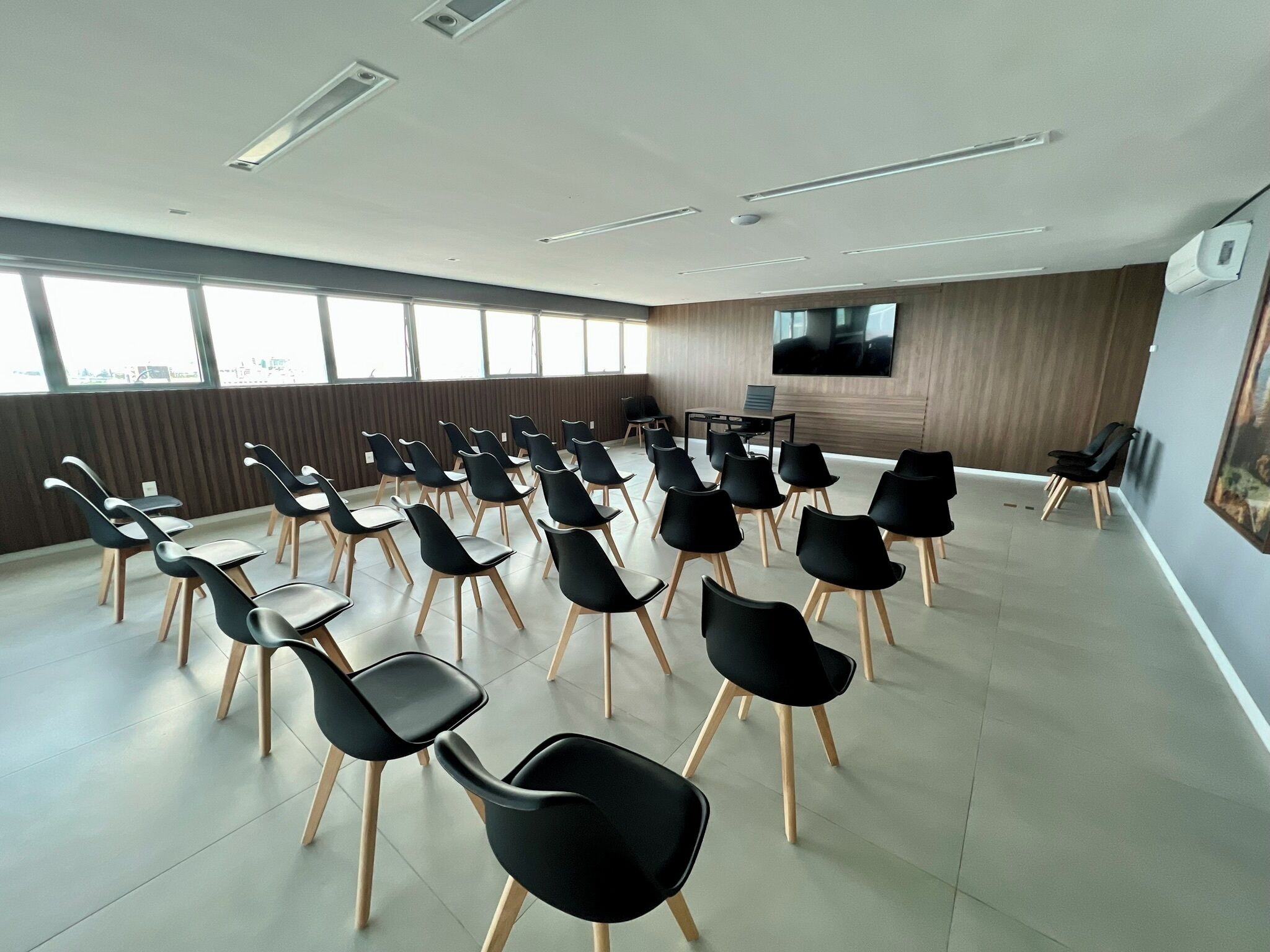 A conference room with rows of black chairs and tables, large windows, a flat-screen TV, and a wooden panel wall.