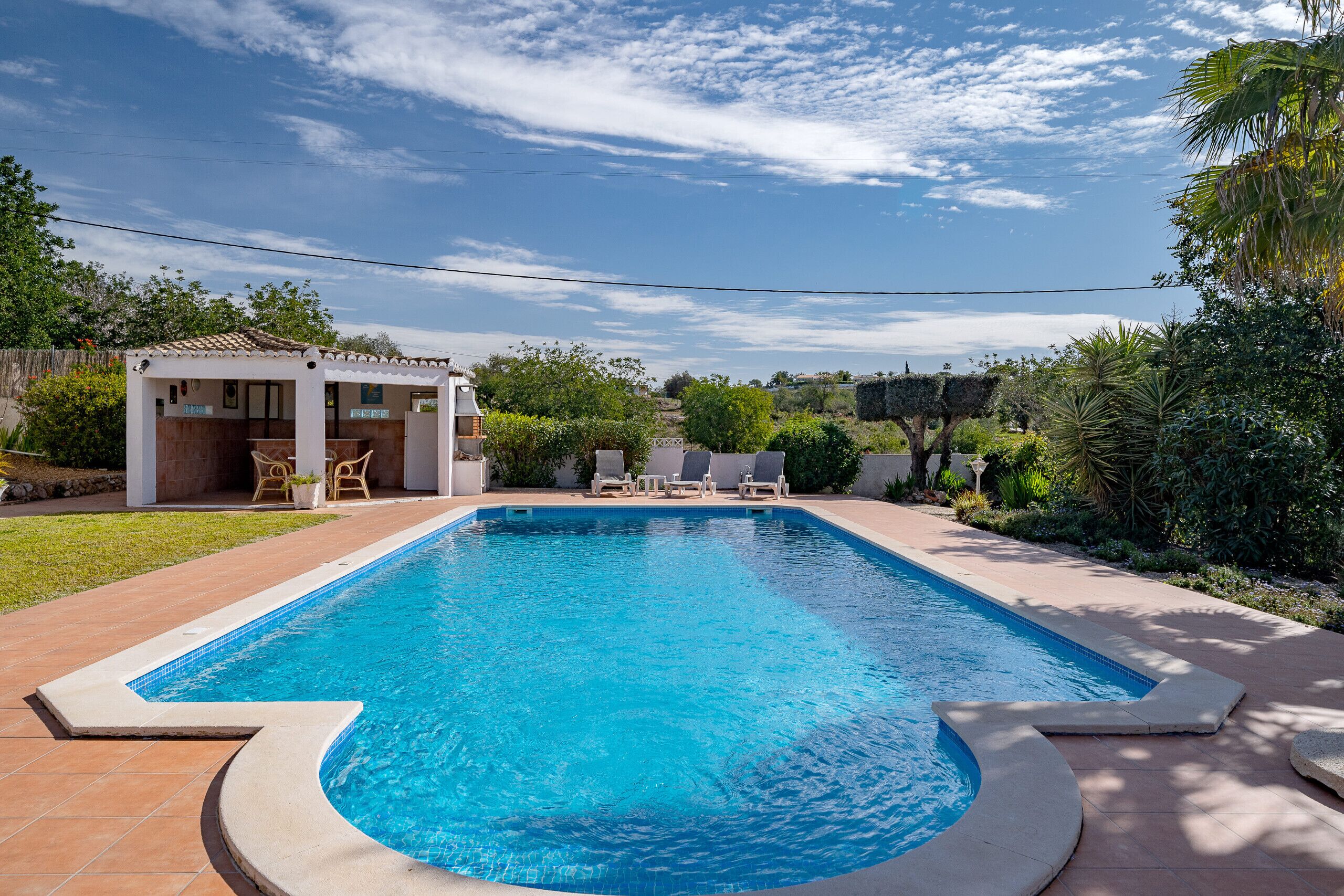 A rectangular swimming pool with a tiled deck, surrounded by greenery and a small building with outdoor seating.