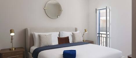 Una cama bien hecha con una toalla azul, un espejo redondo en la pared y una mesita de noche con una lámpara.