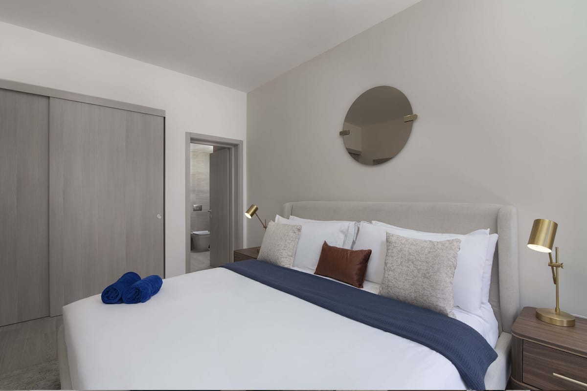 A neatly made bed with a blue blanket, a round mirror on the wall, and a bedside lamp.