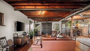 A spacious living area with a leather sofa, wooden flooring, exposed ductwork, and a modern kitchen in the background.
