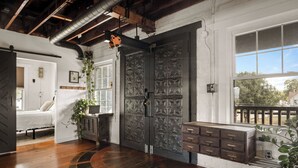 A room with a large, ornate door, a bed, a wooden floor, and a window with a view of trees.