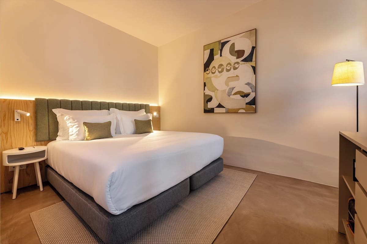 Una habitación de hotel moderna con una cama grande, una mesita de noche, una lámpara y un cuadro abstracto colgado en la pared.