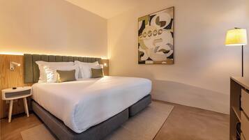 A modern hotel room with a large bed, a bedside table, a lamp, and a wall-mounted abstract painting.