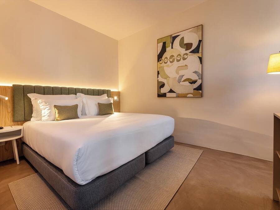 A modern hotel room with a large bed, a bedside table, a lamp, and a wall-mounted abstract painting.