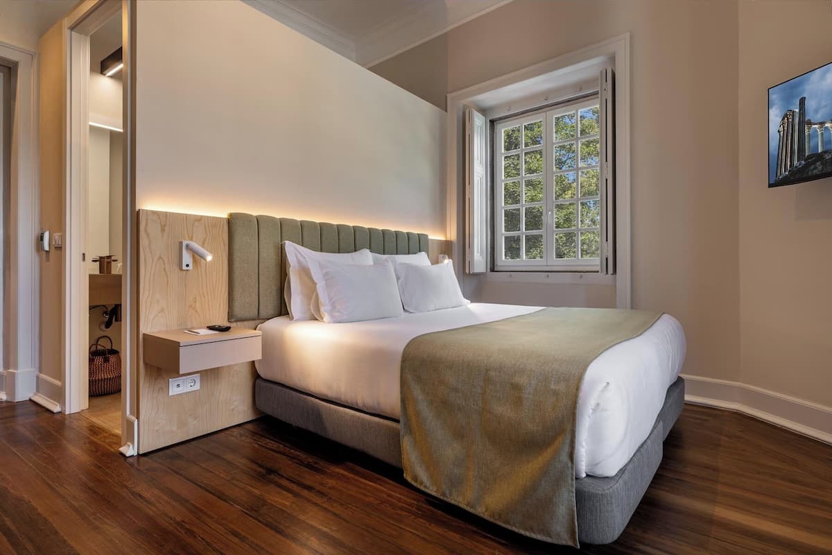 Un dormitorio con cama, mesita de noche, lámpara y ventana con vista a árboles.