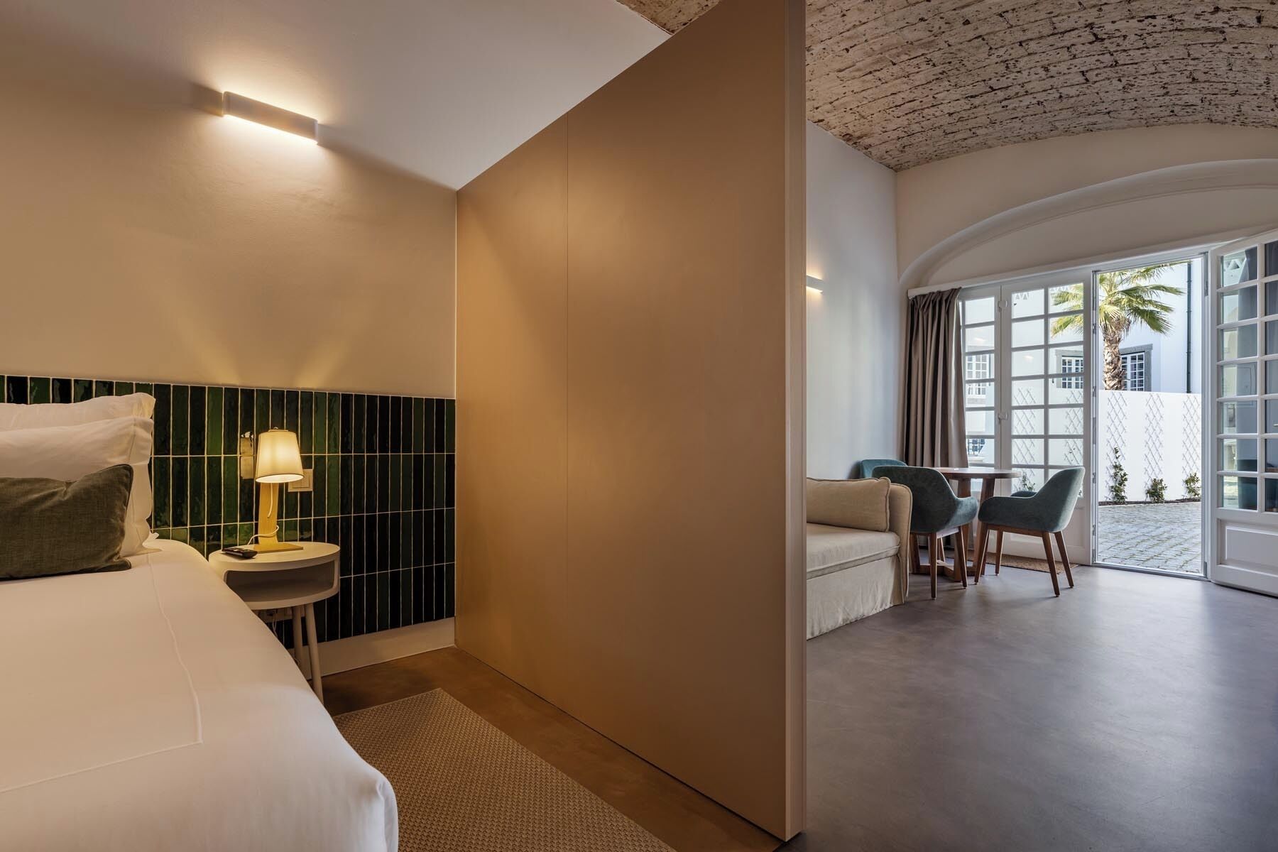 A hotel room with a bed, bedside table, lamp, and a view of the outdoor seating area.