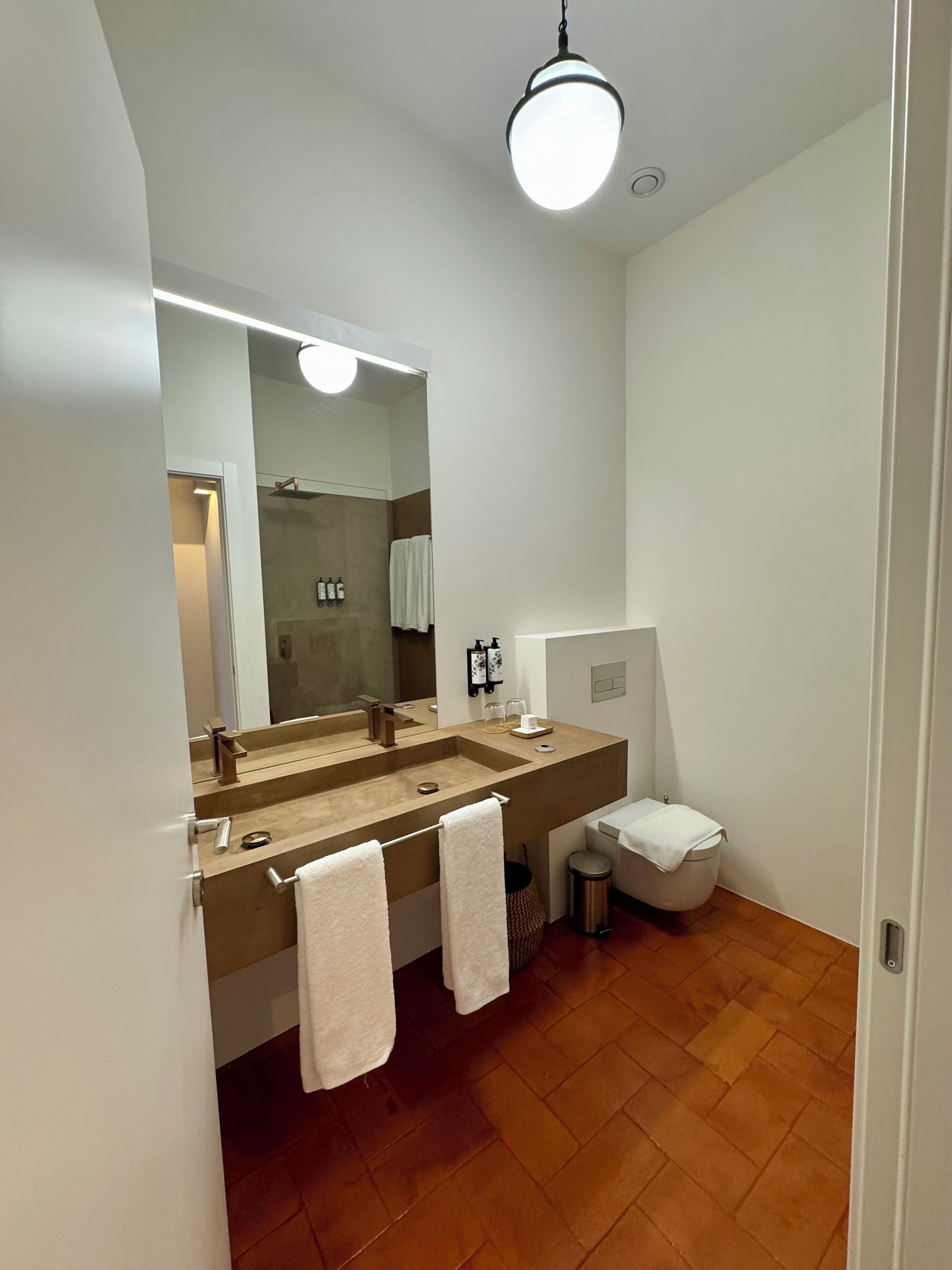 A modern bathroom with a large mirror, two sinks, a toilet, and a trash bin.