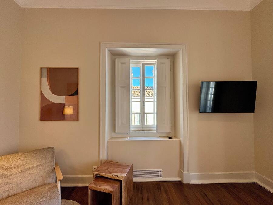 Una habitació amb finestra, televisió de pantalla plana penjada a la paret i un petit tamboret de fusta.