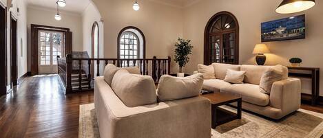 Un salón amplio con un sofá beige grande, una mesa de centro y una alfombra. Hay ventanas de arco y una escalera que conduce a otro nivel.