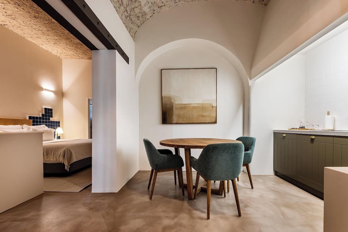 Una cocina moderna con una mesa redonda de madera y cuatro sillas, un comedor con techo alto abovedado, y una cama visible en la habitación contigua.
