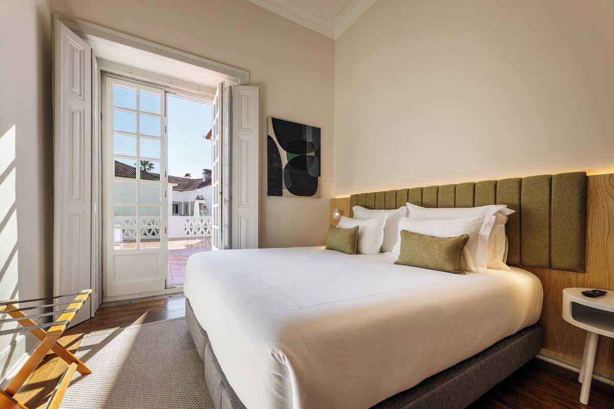 Habitación de hotel con una cama grande, cabecera de madera, una mesita de noche pequeña y vistas a un área exterior a través de una puerta abierta.