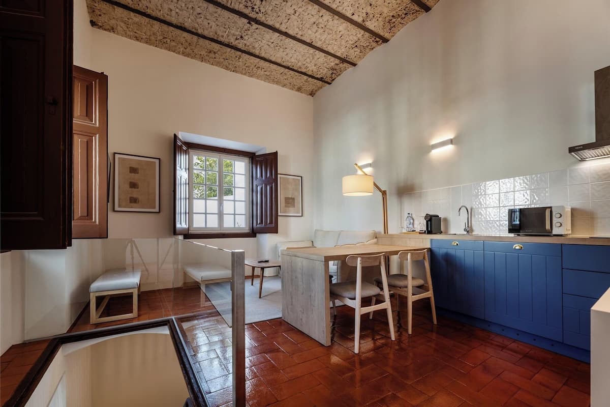Una cocina compacta con una mesa de madera, armarios azules y una ventana con contraventanas abiertas.