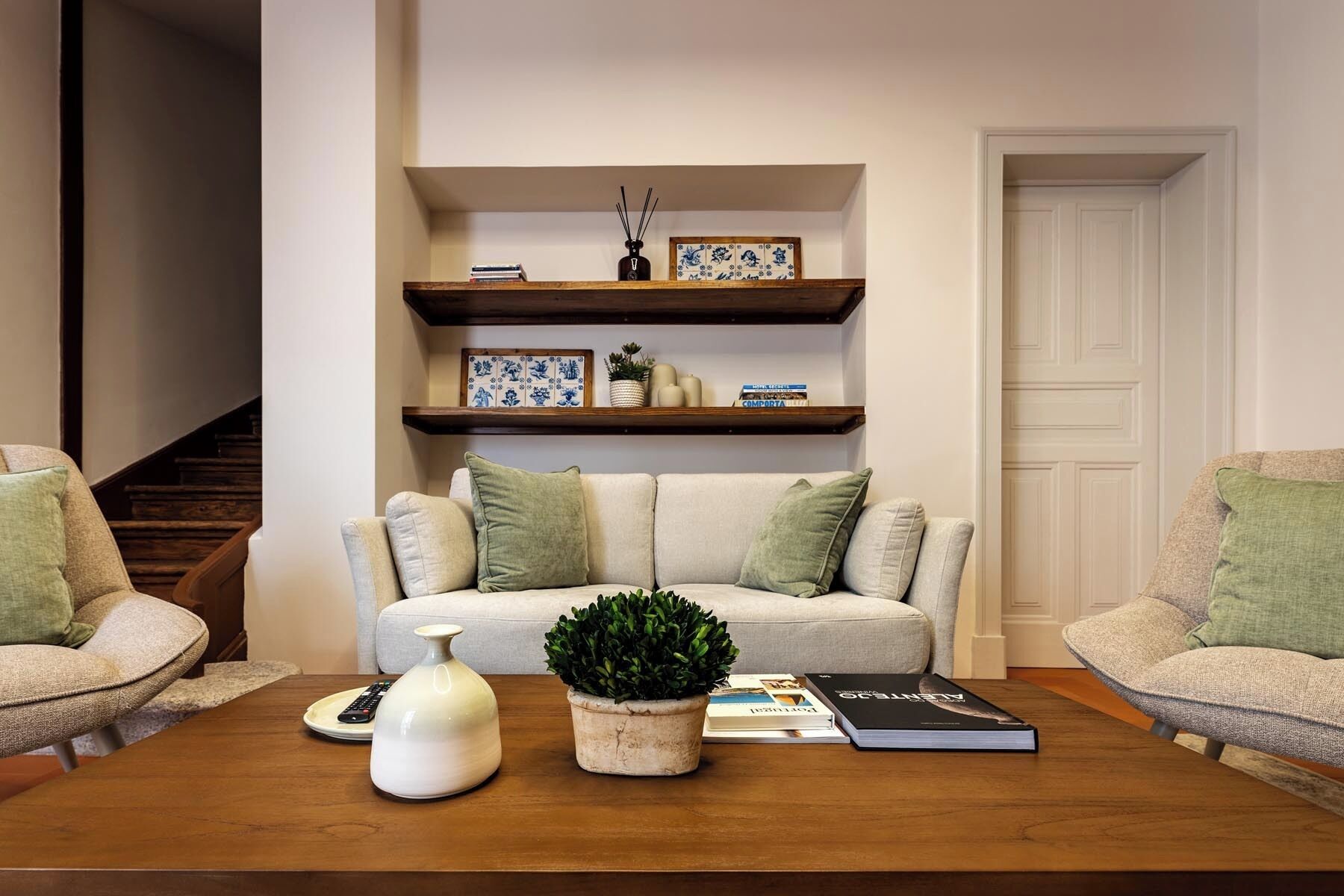 Ein Wohnzimmer mit einer Couch, zwei Sesseln, einem Couchtisch und Dekoration. Über der Couch befinden sich zwei Holzregale mit Büchern und dekorativen Objekten.