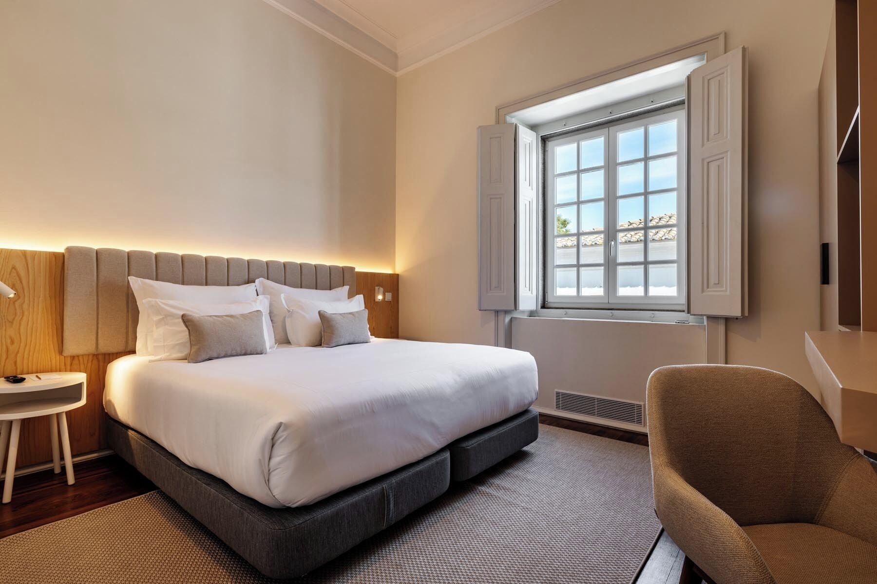 A modern hotel room with a large bed, a bedside table, a chair, and a window with a view.