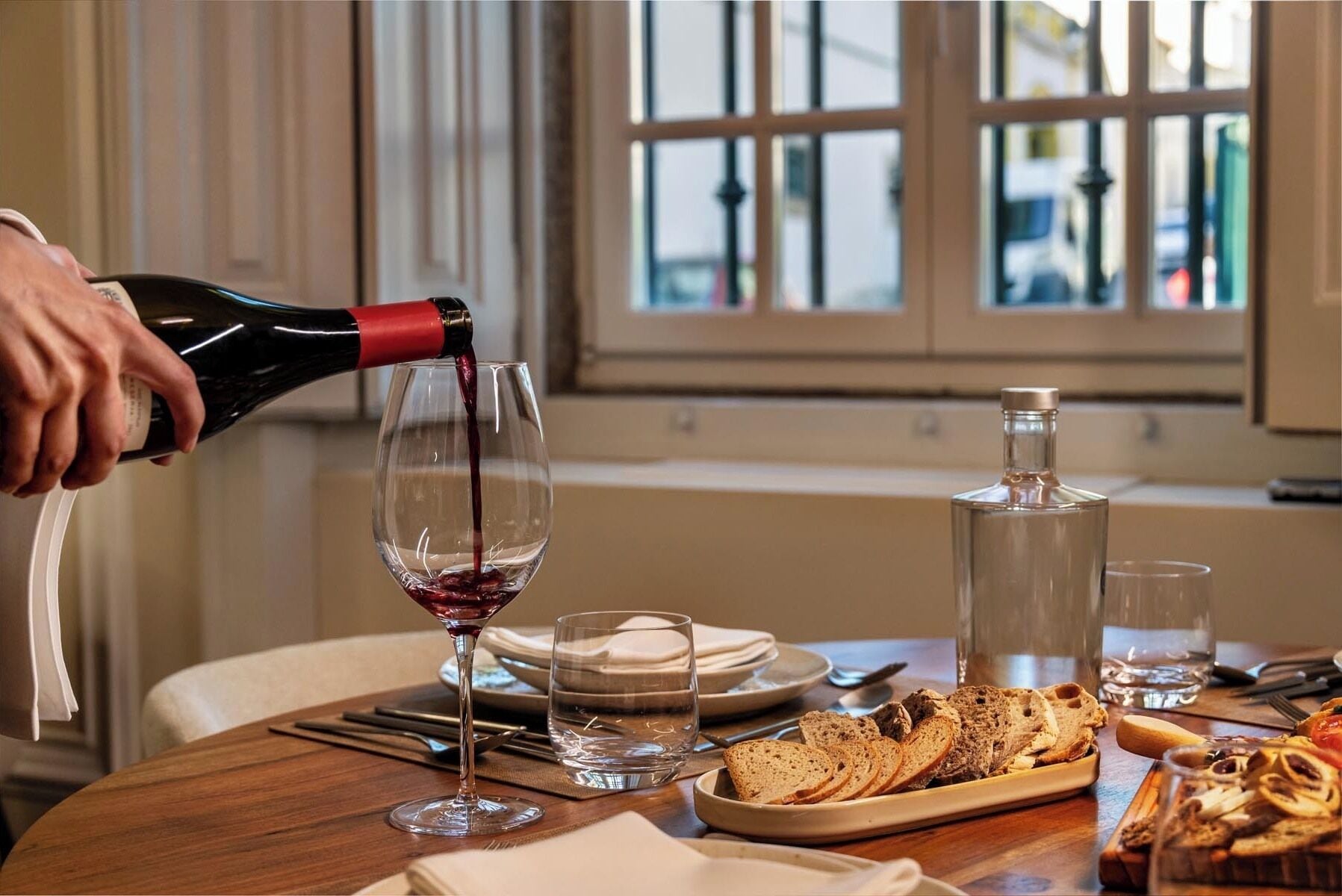 A dining table with a bottle of red wine being poured into a glass, a bread basket, and a bottle of water.