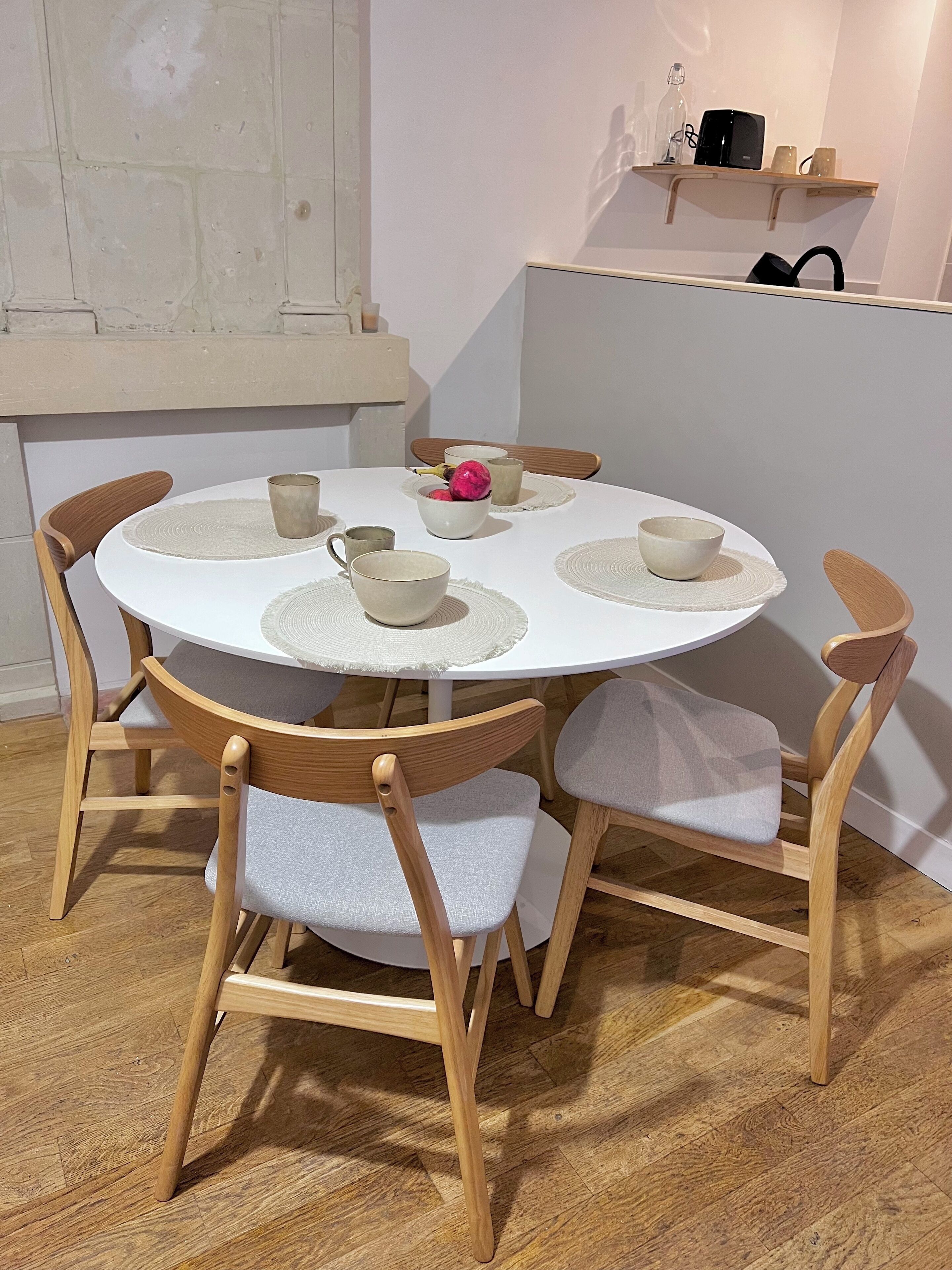 Una mesa redonda de comedor con cuatro sillas, cada una con estructura de madera y asientos acolchados grises. La mesa está rodeada de individuales y tiene varias tazas de cerámica y un bol con elementos rosas.
