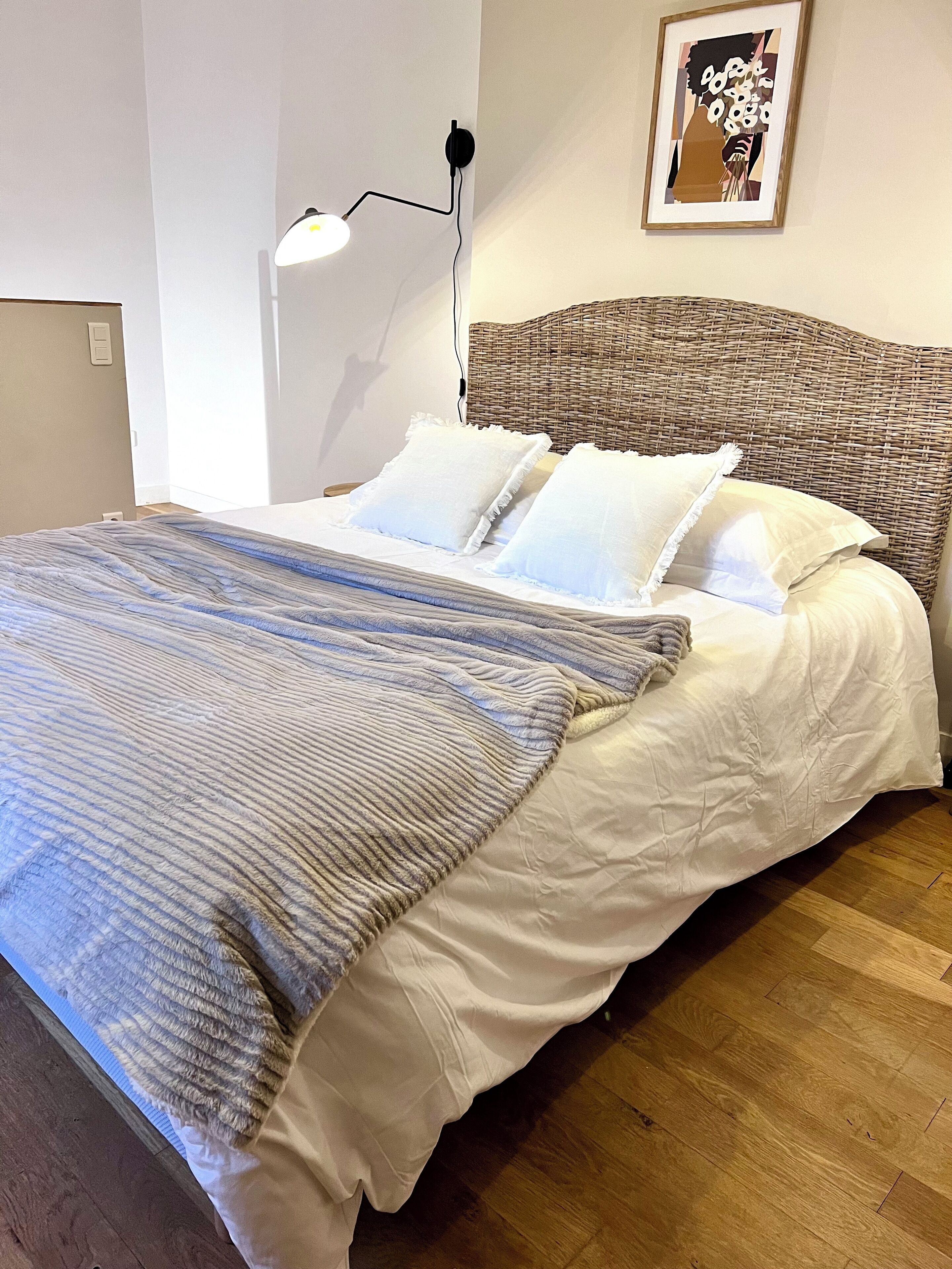 Un lit bien fait, avec une couverture à rayures, une tête de lit en osier et un tableau encadré accroché au mur.