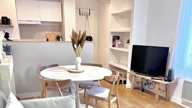 A modern living room with a sofa, a dining table with chairs, a TV on a stand, and a kitchen area in the background. - Jeanne's apartment. 500 from the castle, in a quiet, green setting (Chinon)
