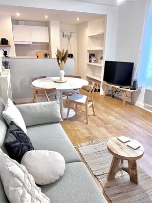 Ein modernes Wohnzimmer mit einer Couch, einem Esstisch mit Stühlen, einem Fernseher auf einem Schrank und einer Küche im Hintergrund.