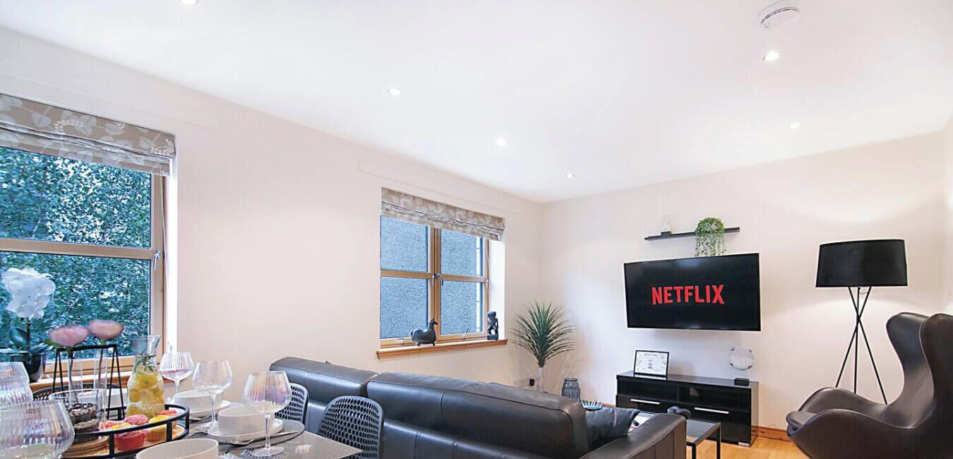 Una sala de estar moderna con un sofá de cuero negro, una mesa de comedor con cubierta de vidrio, un ventanal con vista a árboles, un televisor de pantalla plana montado en la pared y una lámpara de pie.