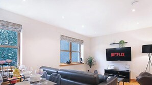 Una sala de estar moderna con un sofá de cuero negro, una mesa de comedor con cubierta de vidrio, un ventanal con vista a árboles, un televisor de pantalla plana montado en la pared y una lámpara de pie.
