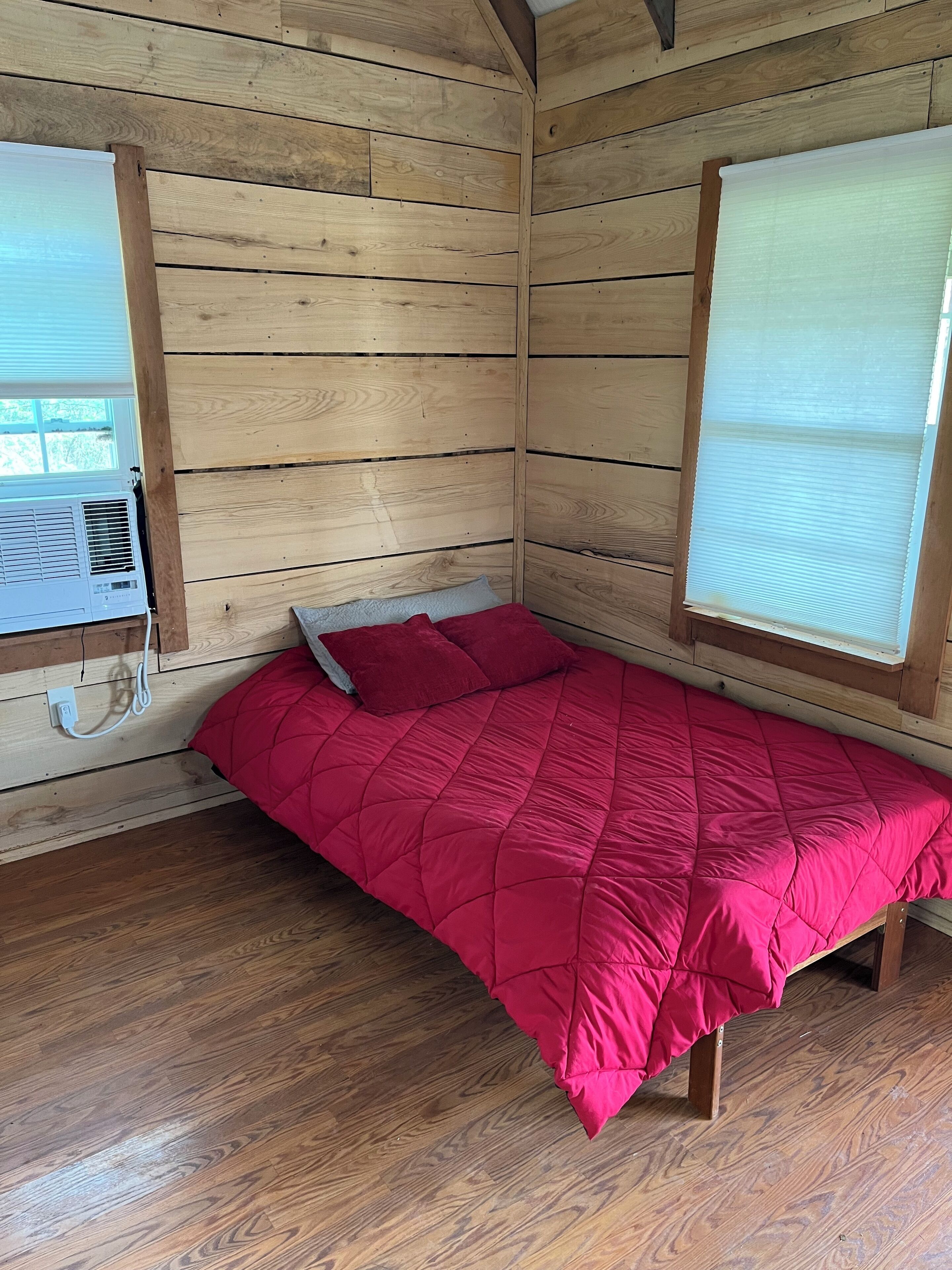 Um quarto pequeno de madeira com uma cama, uma janela com persianas e um aparelho de ar condicionado.