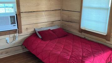 Um quarto pequeno de madeira com uma cama, uma janela com persianas e um aparelho de ar condicionado.