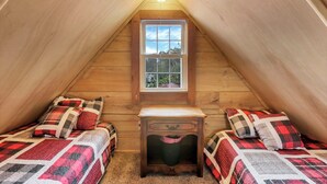 A cozy attic bedroom with wooden walls, a window with a view, two beds with checkered bedding, and a small bedside table. - Cozy Cabin (New Lexington)