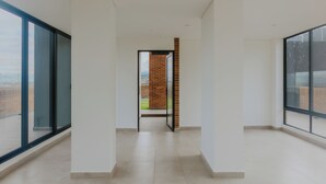 Een modern, ruim interieur met grote ramen, een centrale open doorgang naar een bakstenen muur, en een betegelde vloer.