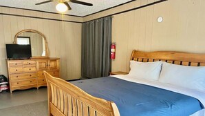 Un dormitorio con una cama de madera, un televisor sobre un aparador de madera, un ventilador de techo y un extintor.
