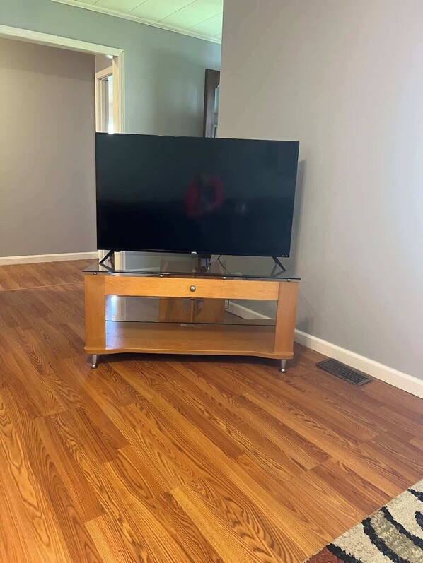 A flat-screen TV on a wooden stand with a drawer, placed on a wooden floor in a room with a plain wall and a door in the background.