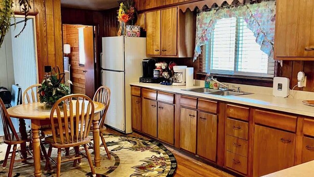Cocina compacta con armarios de madera, una mesa de comedor con sillas, un refrigerador, una cafetera y una ventana con cortina decorativa.
