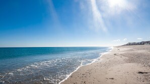 Ein Strand mit klarem, blauem Wasser, einem sandigen Ufer und einer Reihe von Häusern im Hintergrund.