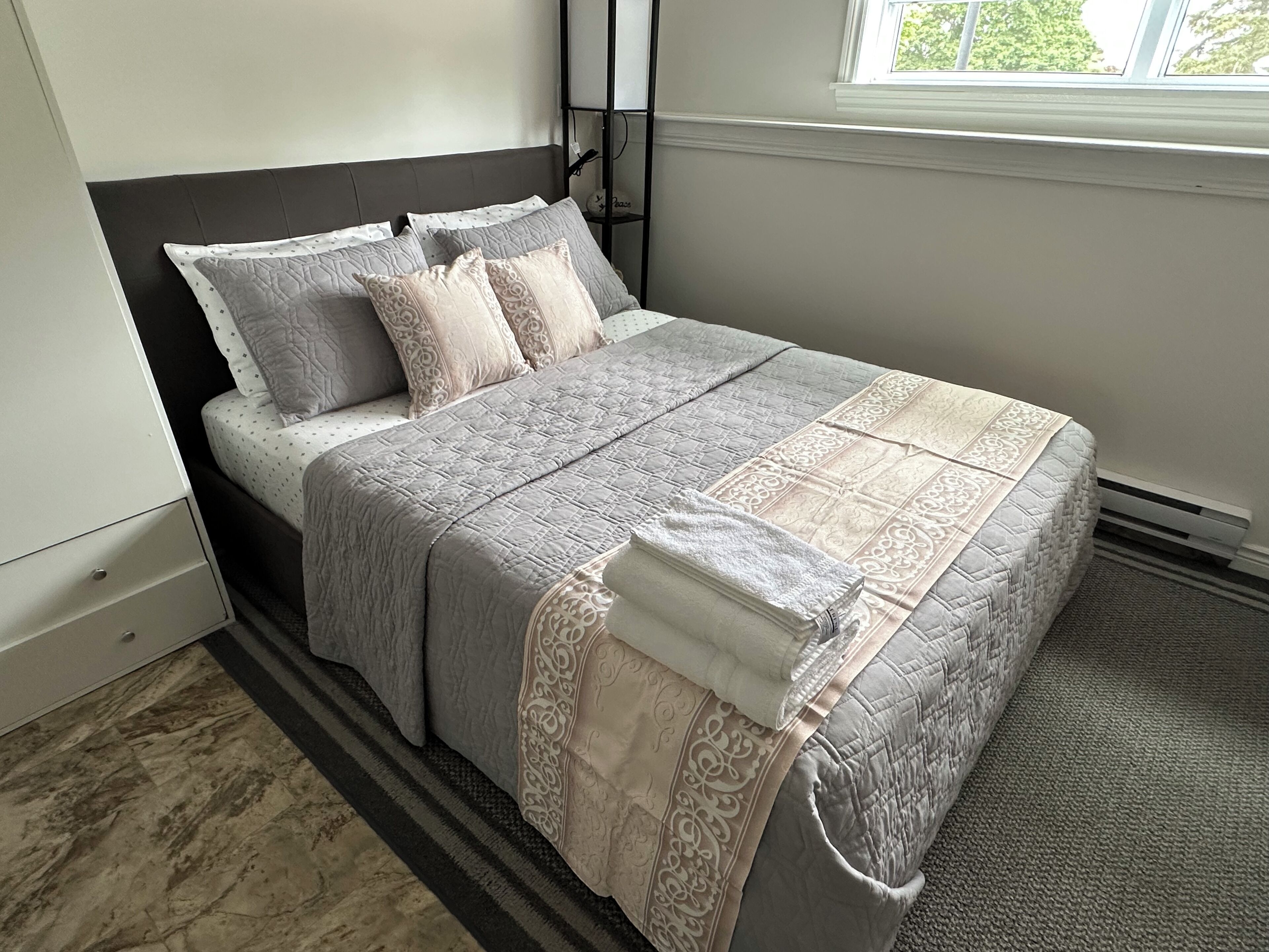 A neatly made bed with a grey and white quilt, a black headboard, and a window with a view of trees.