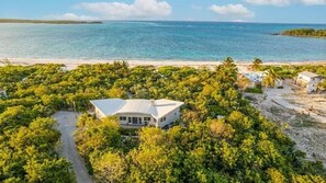 Un bâtiment blanc entouré d’une végétation tropicale luxuriante, avec une plage et l’océan en arrière-plan.