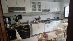 A compact kitchen with white cabinets, a gas stove, and a coffee maker. There is a dining area with a round table and white wicker chairs. - Casa SantaMaria (Lousame)