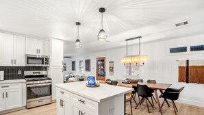 Een moderne keuken met witte kastjes, roestvrijstalen apparatuur en een centraal keukenblok. Er is een eethoek met een houten tafel en zwarte stoelen.