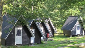 A row of small, dark wooden cabins with pitched roofs, surrounded by lush greenery and a gravel path. - Holiday Home for 2 in Stenungsund (Stenungsund)