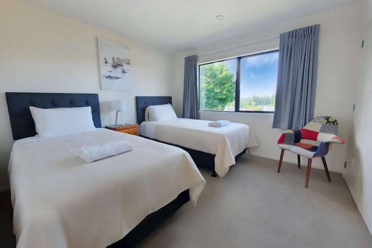 Ein Hotelzimmer mit zwei Betten, einem fensterbelaubten Fenster, einem Sessel und einem Bild an der Wand.