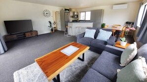 Ein modernes Wohnzimmer mit grauem Sofa, einem Holztisch, einem Flachbildfernseher, einer Küchenzeile mit Barhocker und einem Esstisch mit Stühlen.