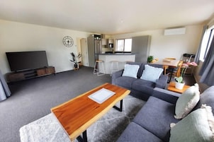 Una sala de estar moderna con un sofá gris, una mesa de centro de madera, un televisor de pantalla plana, una zona de cocina con taburetes altos y una mesa de comedor con sillas.