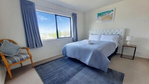 Ein Schlafzimmer mit Bett, Stuhl, Nachttisch, einem Fenster mit Vorhängen und einem Bild an der Wand.